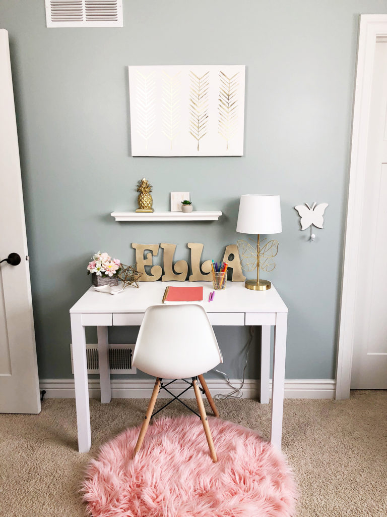 White desk with butterfly lamp, white chair, pink rug and flowers. 